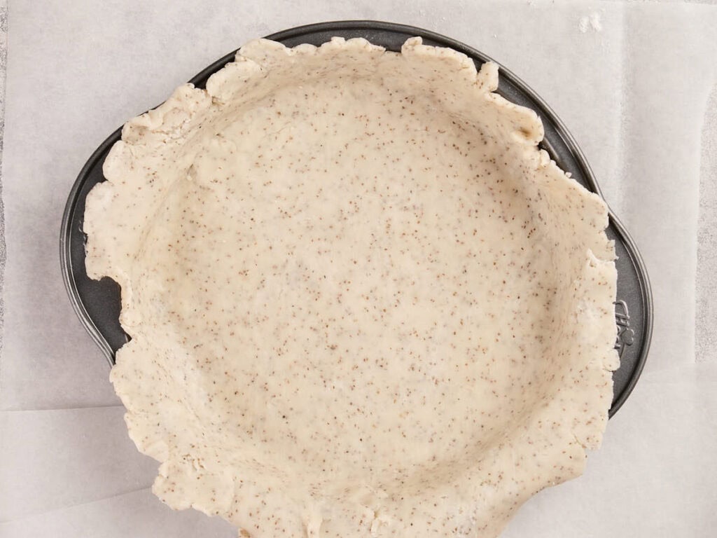 Unbaked pie crust dough is pressed into a metal pie pan on a sheet of parchment paper.