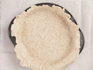 Unbaked pie crust dough is pressed into a metal pie pan on a sheet of parchment paper.
