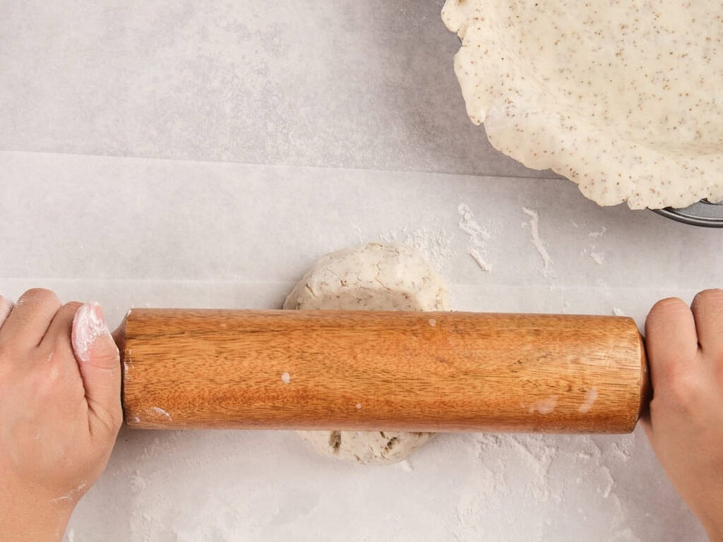 Two hands roll out dough with a wooden rolling pin on a floured surface.