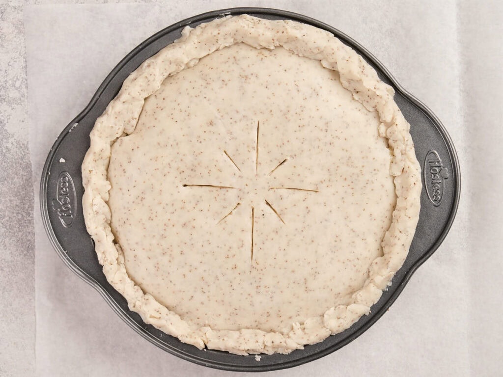 Unbaked pie with a crimped, speckled crust and eight slits cut in the center sits in a round metal pie pan.