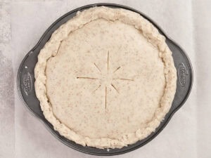 Unbaked pie with a crimped, speckled crust and eight slits cut in the center sits in a round metal pie pan.