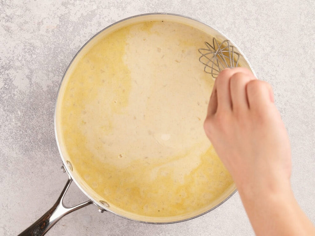 A hand whisking a creamy yellow sauce in a metal pan on a light gray surface.