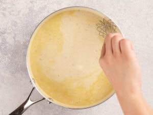 A hand whisking a creamy yellow sauce in a metal pan on a light gray surface.