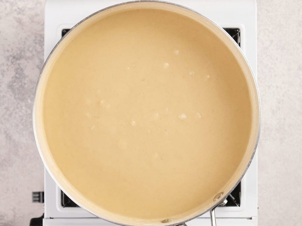 A pot of light beige, creamy sauce is heating on a stovetop burner, with small bubbles forming on the surface.