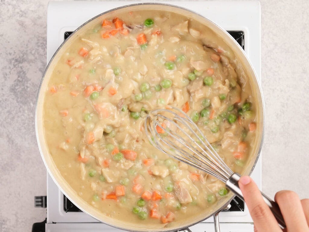 A hand uses a whisk to stir a creamy mixture of carrots, peas, and chicken in a pan on a stovetop.