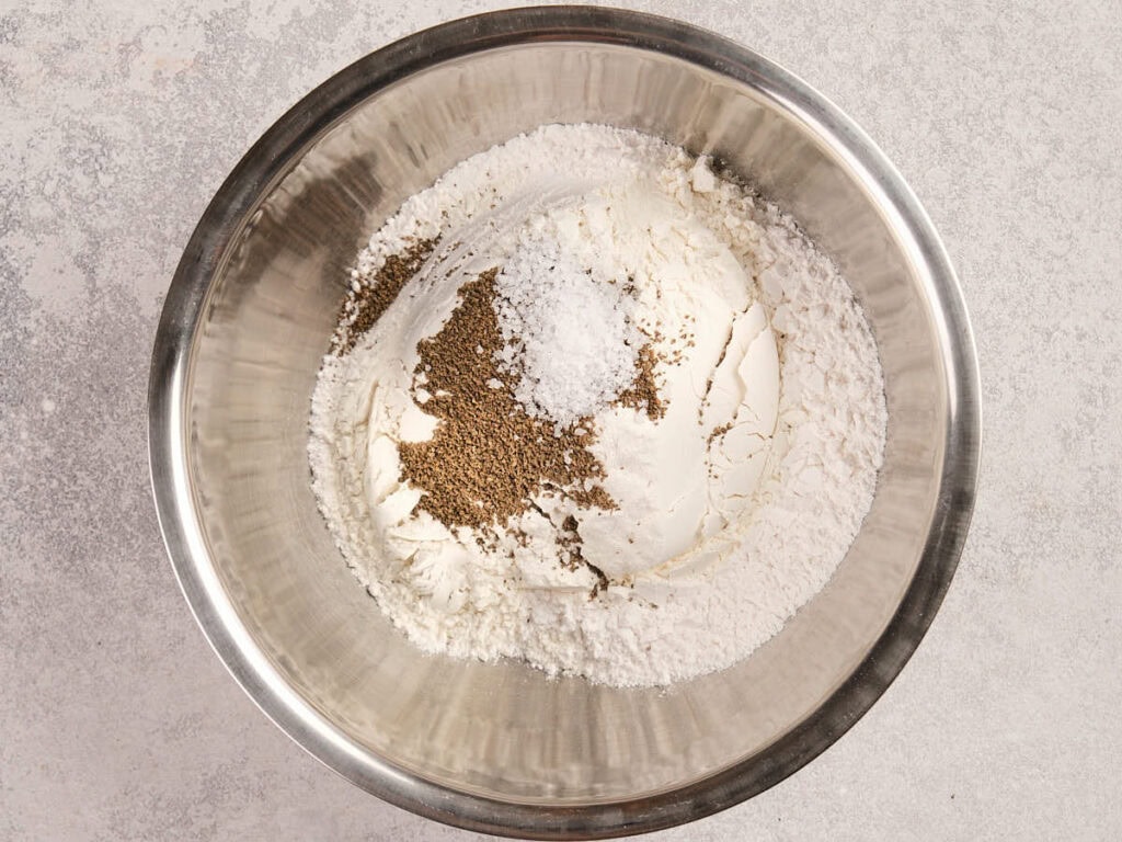 A metal mixing bowl containing flour, celery seed and salt on a light countertop.
