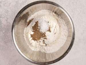 A metal mixing bowl containing flour, celery seed and salt on a light countertop.