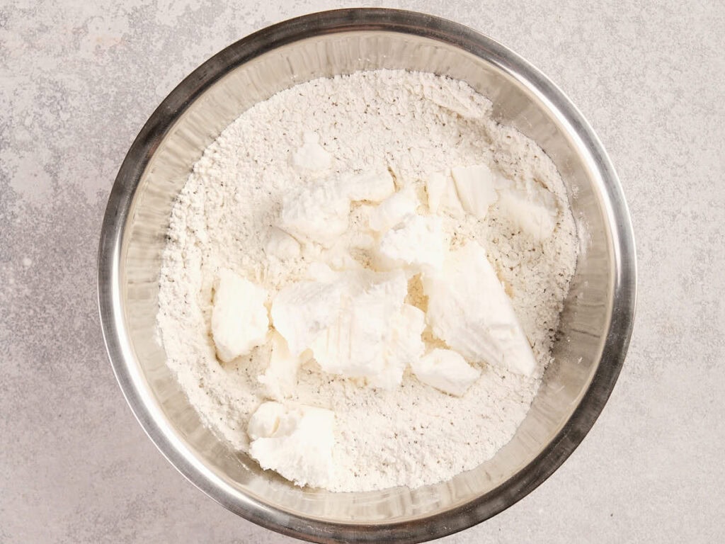 A metal bowl containing flour with chunks of lard on a light-colored surface.