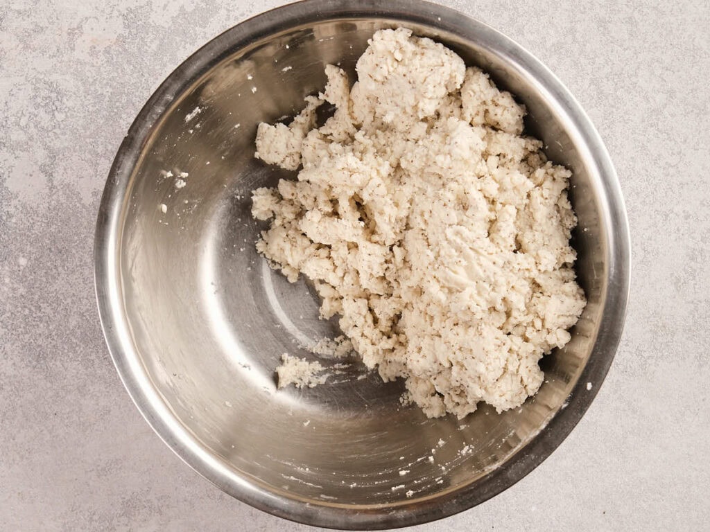 A stainless steel bowl containing a mixture of crumbly, white dough sits on a light-colored countertop.