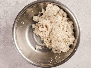 A stainless steel bowl containing a mixture of crumbly, white dough sits on a light-colored countertop.