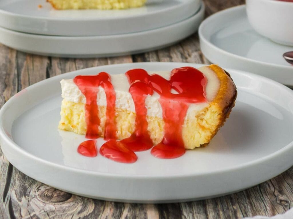 A slice of cheesecake topped with red strawberry sauce on a white plate, with another plate and bowl in the background on a wooden surface.