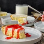 A slice of cheesecake with red sauce on a white plate, with a fork holding a bite and a glass of milk in the background.