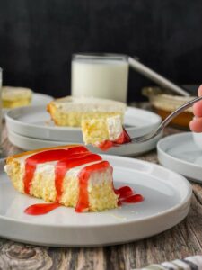A slice of cheesecake with red sauce on a white plate, with a fork holding a bite and a glass of milk in the background.