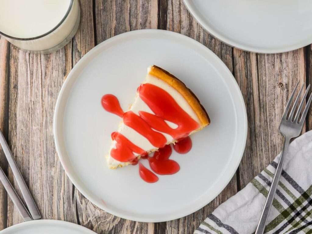 A slice of cheesecake with red strawberry sauce on a white plate, set on a wooden table with a fork, napkin, and a glass of milk nearby.