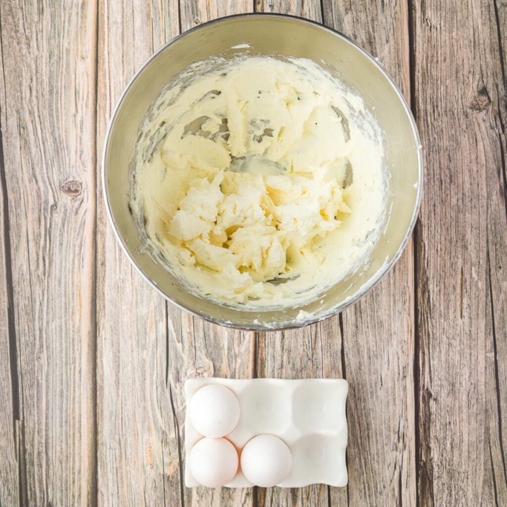 A metal mixing bowl with creamed butter sits above a ceramic tray holding three eggs on a wooden surface.