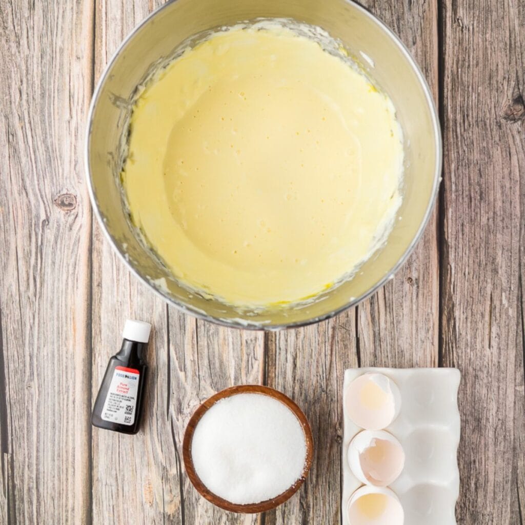 A mixing bowl with yellow batter, a bottle of vanilla extract, a bowl of sugar, and cracked eggs in a tray on a wooden surface.