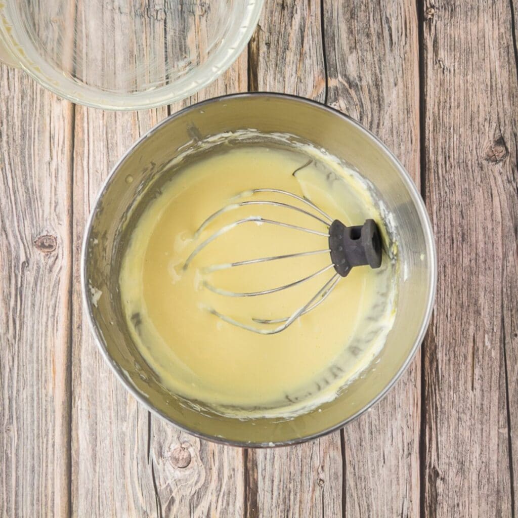 A metal mixing bowl with a whisk sits on a wooden surface, containing a smooth, pale yellow batter or mixture.