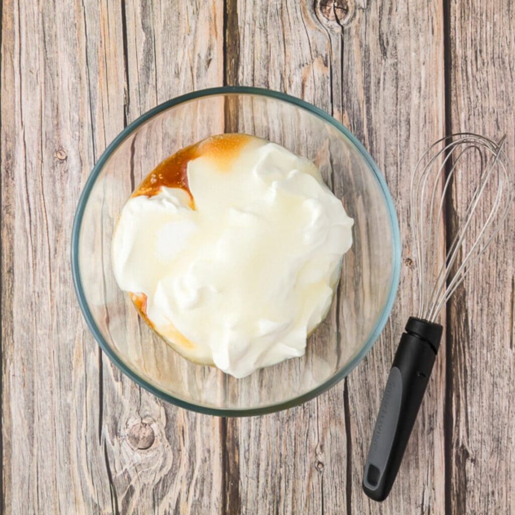 A glass bowl with whipped egg whites and a caramel-colored liquid on a wooden surface next to a metal whisk with a black handle.