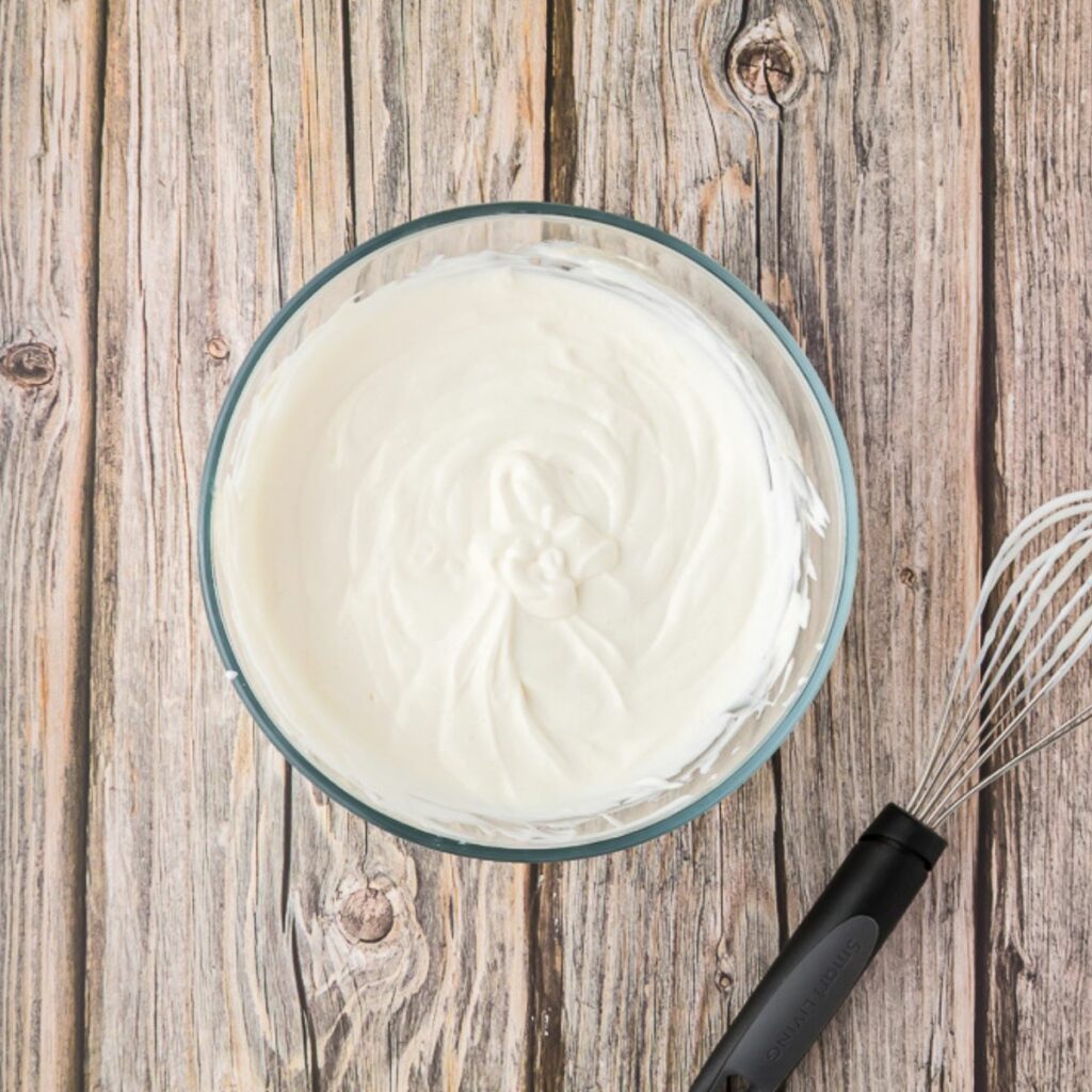 A glass bowl of whipped cream sits on a wooden surface next to a metal whisk with a black handle.