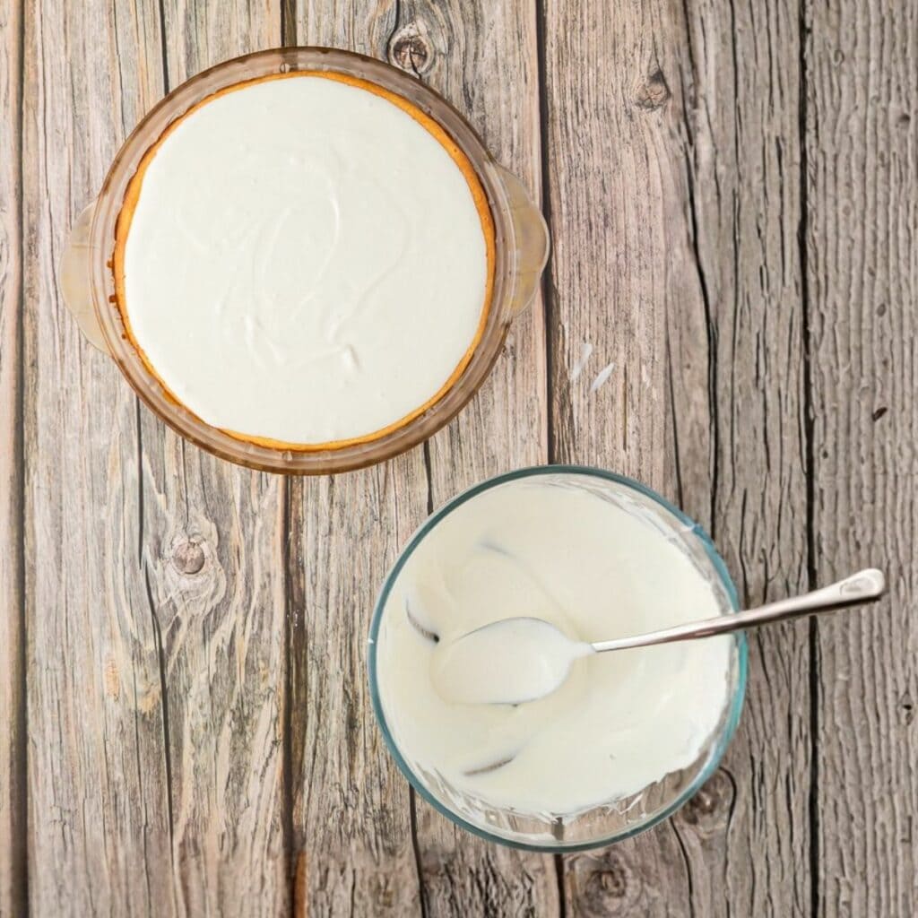 A glass pie dish with a cheesecake topped with a white layer sits next to a glass bowl of white mixture and a spoon on a wooden surface.