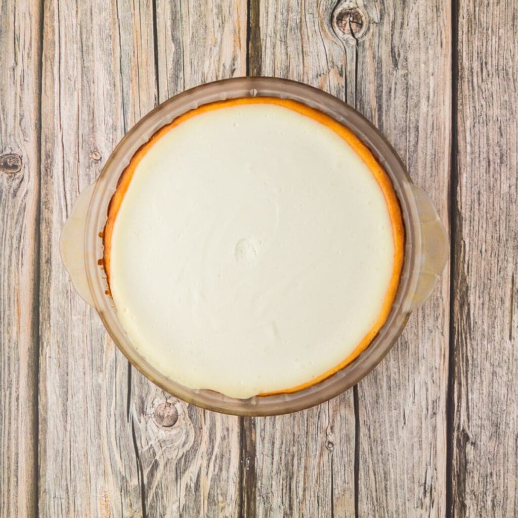 A round cheesecake with a smooth, creamy top in a glass dish, placed on a rustic wooden surface.