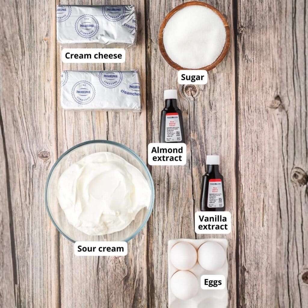 Cheesecake ingredients on a wooden surface: cream cheese, sugar, almond extract, vanilla extract, sour cream, and eggs.