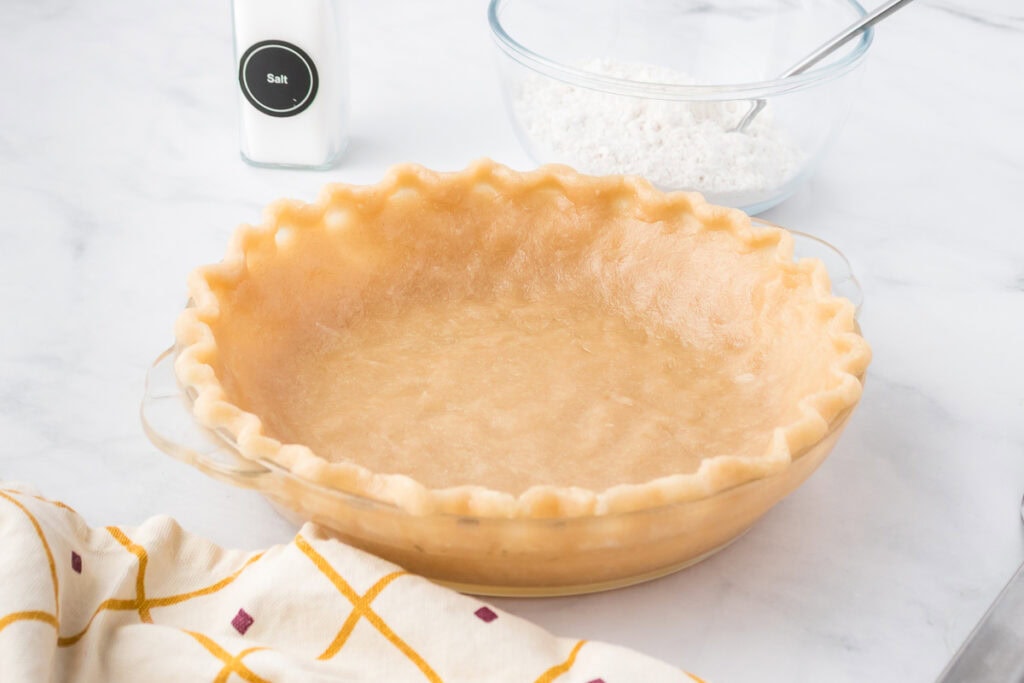 An unbaked pie crust in a glass pie dish sits on a countertop, with a bowl of flour, a container of salt, and a patterned towel nearby.