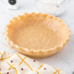 Unbaked pie crust in a glass pie dish with a fluted edge, next to a bowl of flour with a spoon, a salt container, and a patterned kitchen towel.