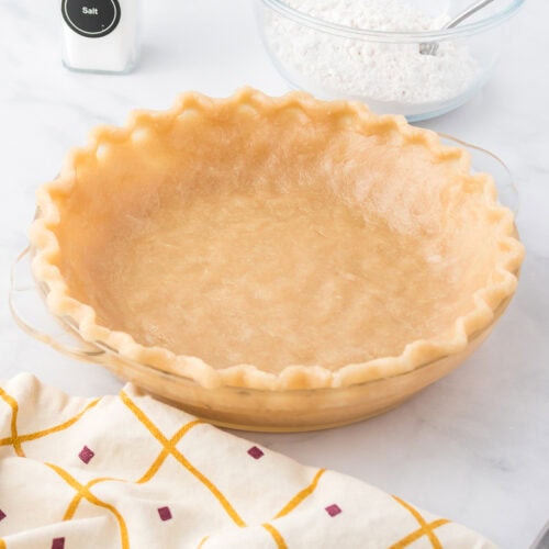 Unbaked pie crust in a glass pie dish with a fluted edge, next to a bowl of flour with a spoon, a salt container, and a patterned kitchen towel.