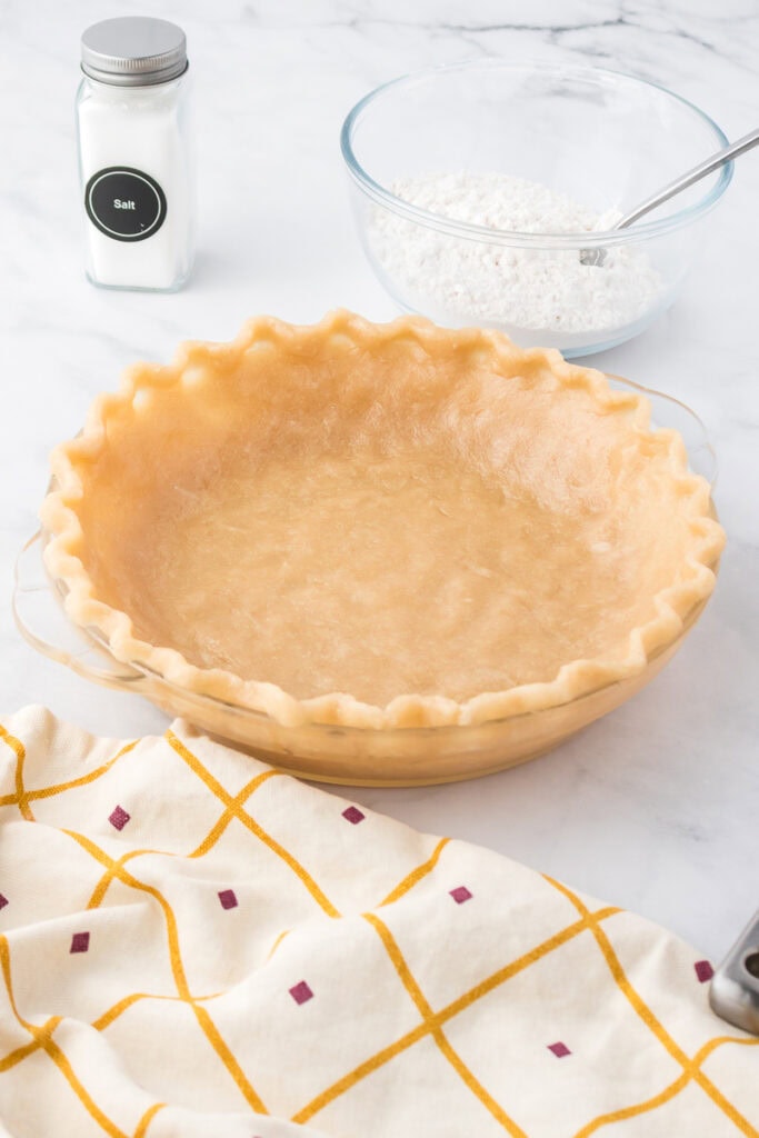 Unbaked pie crust in a glass pie dish with a fluted edge, next to a bowl of flour with a spoon, a salt container, and a patterned kitchen towel.