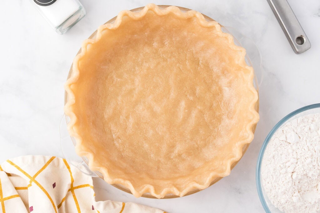 An unbaked pie crust in a glass pie dish on a marble surface, with a bowl of flour, a salt shaker, and a kitchen towel nearby.