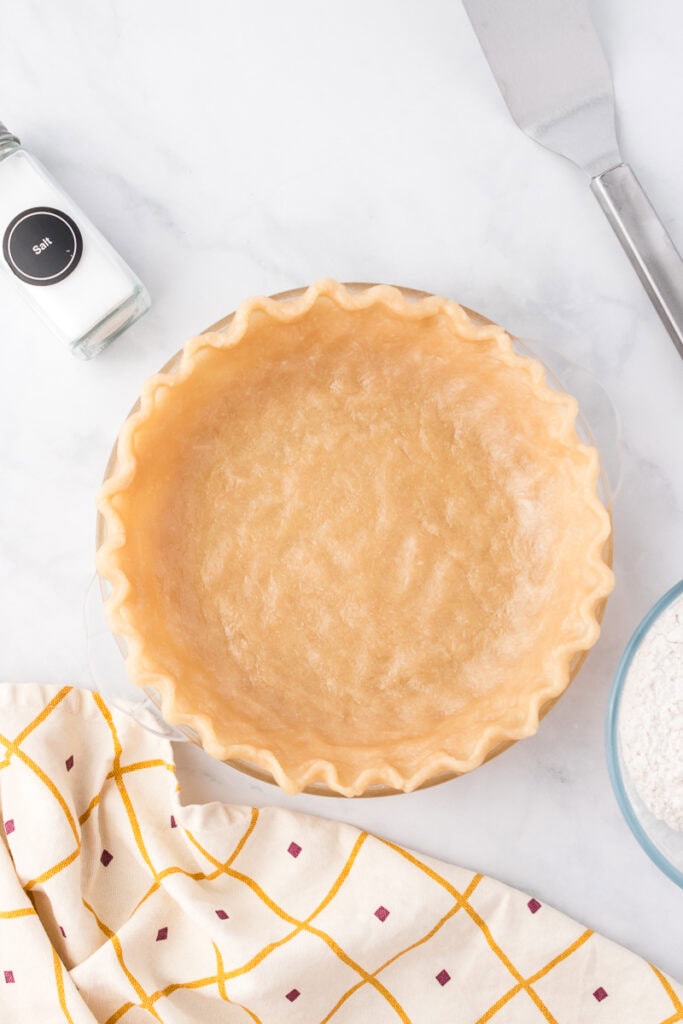 Unbaked pie crust in a glass pie dish on a marble surface, with a salt shaker, spatula, bowl of flour, and patterned towel nearby.