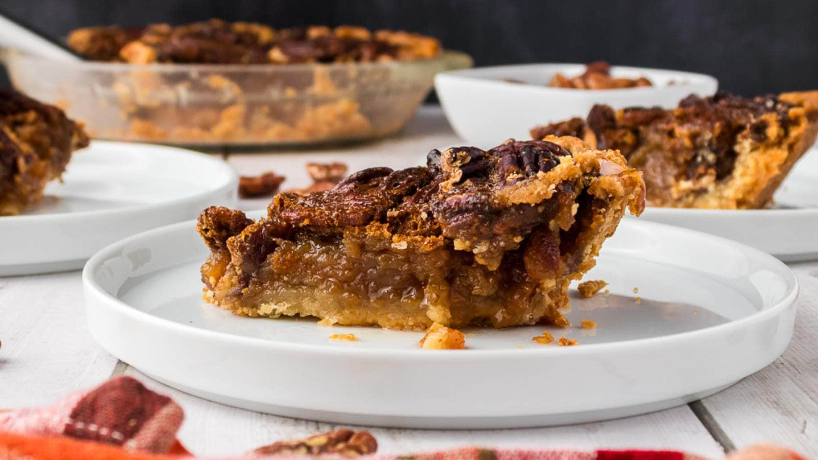 A slice of pecan pie sits on a white plate, with more pie slices and a pie dish in the background on a white table.