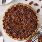 A baked pecan pie in a glass pie dish sits on a white surface, surrounded by pecans, a striped towel, plates, and utensils.