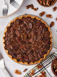 A baked pecan pie in a glass pie dish sits on a white surface, surrounded by pecans, a striped towel, plates, and utensils.