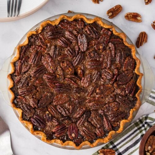 A baked pecan pie in a glass pie dish sits on a white surface, surrounded by pecans, a striped towel, plates, and utensils.