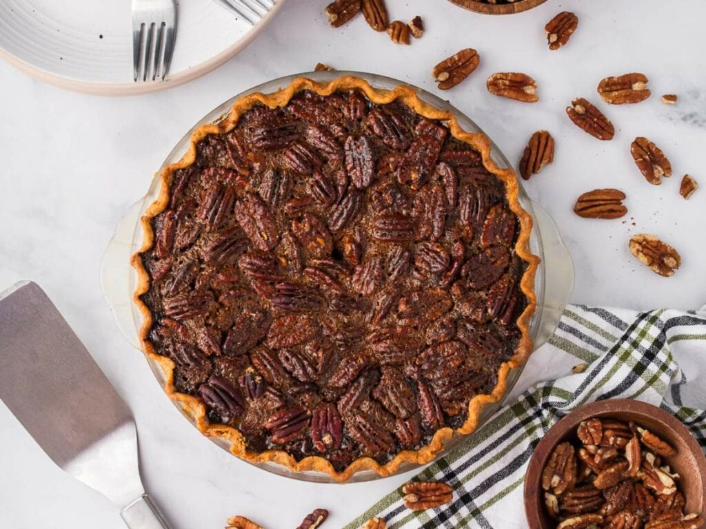 A baked pecan pie in a glass dish on a striped towel, surrounded by pecans, a pie server, and plates with forks.
