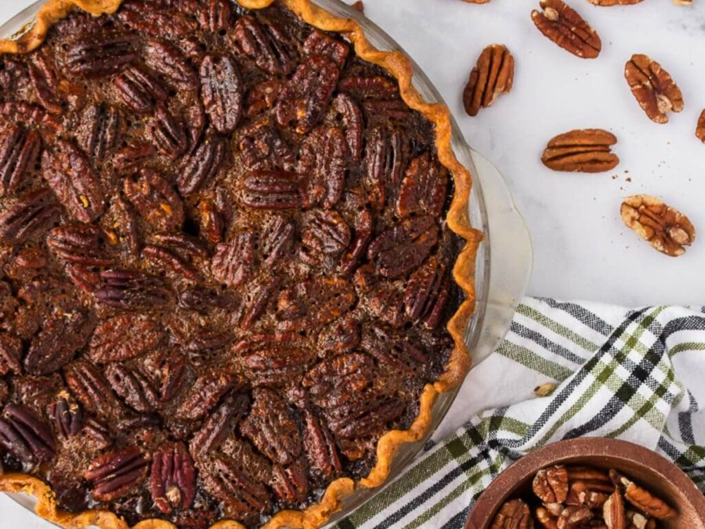 A baked pecan pie in a glass dish, surrounded by scattered pecans on a white surface and a striped kitchen towel.