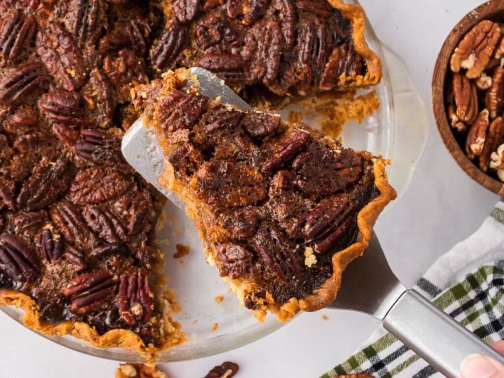 A slice of pecan pie being lifted from a glass pie dish, with whole pecans visible on top and a bowl of pecans nearby.