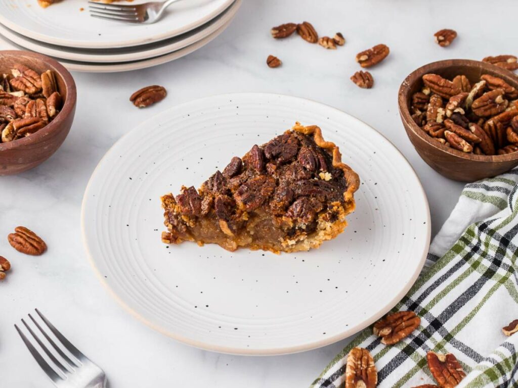 A slice of pecan pie on a white plate, surrounded by pecan nuts in bowls and scattered on a marble surface, with plates, a fork, and a checkered napkin nearby.