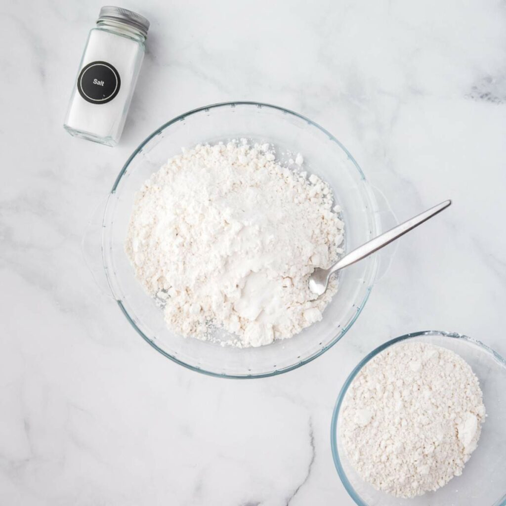 A glass bowl with flour and a spoon, a smaller bowl with more flour, and a jar of salt on a marble surface.