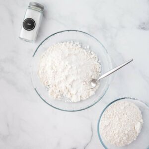 A glass bowl with flour and a spoon, a smaller bowl with more flour, and a jar of salt on a marble surface.