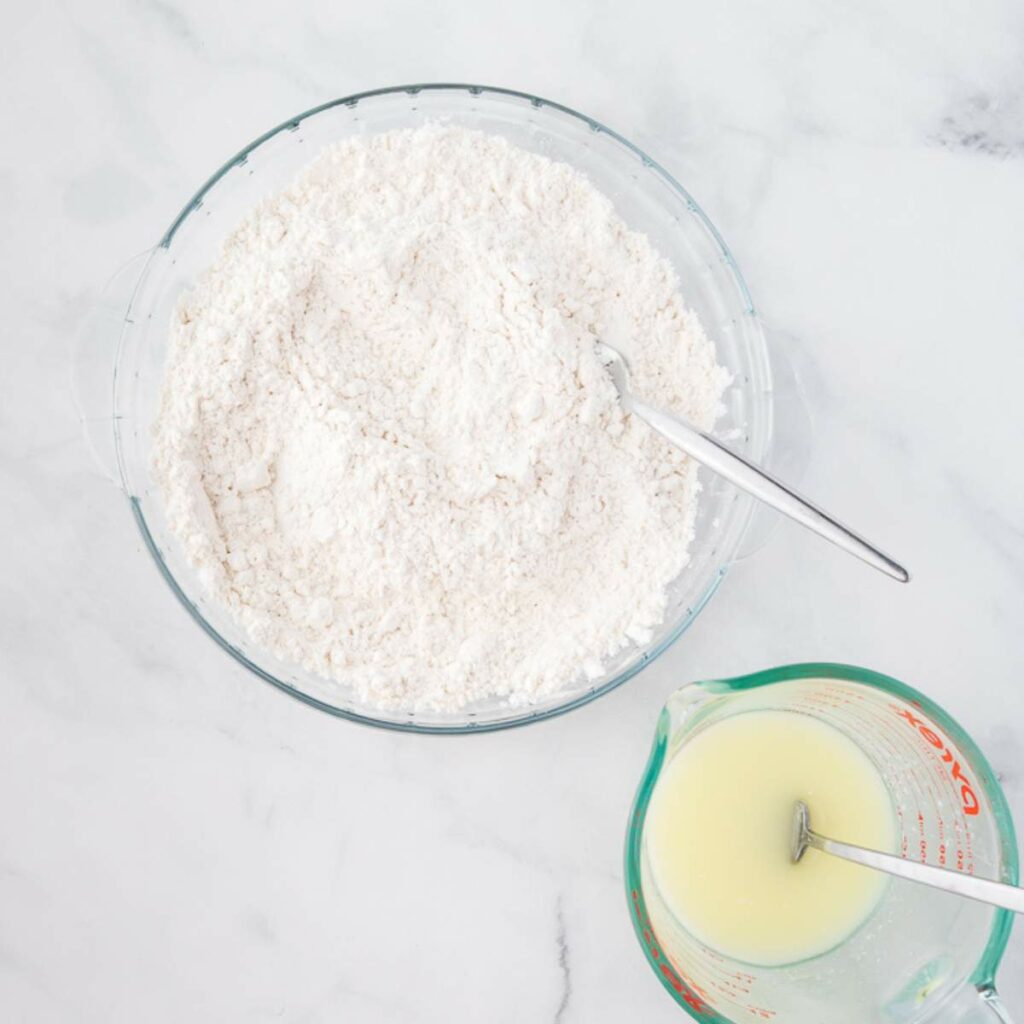 A glass bowl filled with flour mixture and a metal spoon next to a measuring cup containing a yellow liquid with another spoon, both on a white surface.