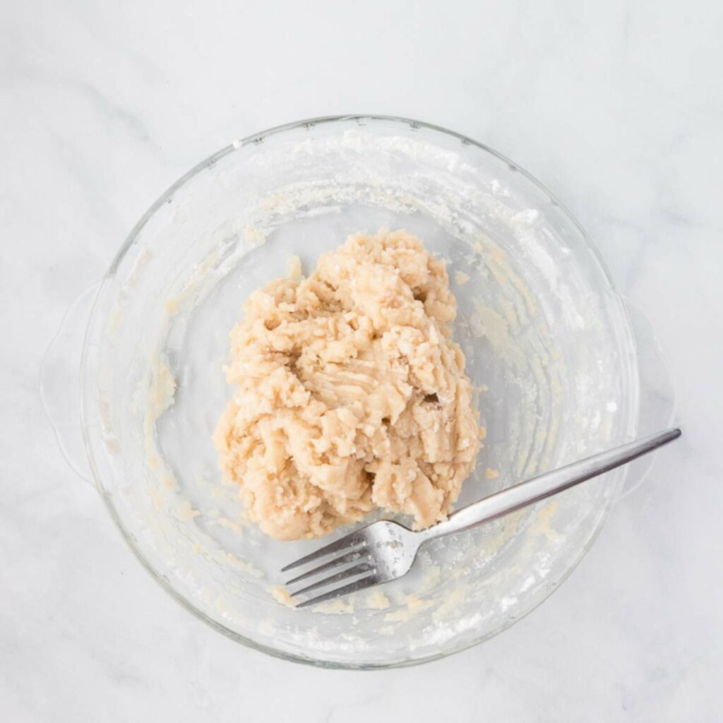 A glass bowl containing a ball of dough with a metal fork resting inside on a white surface.