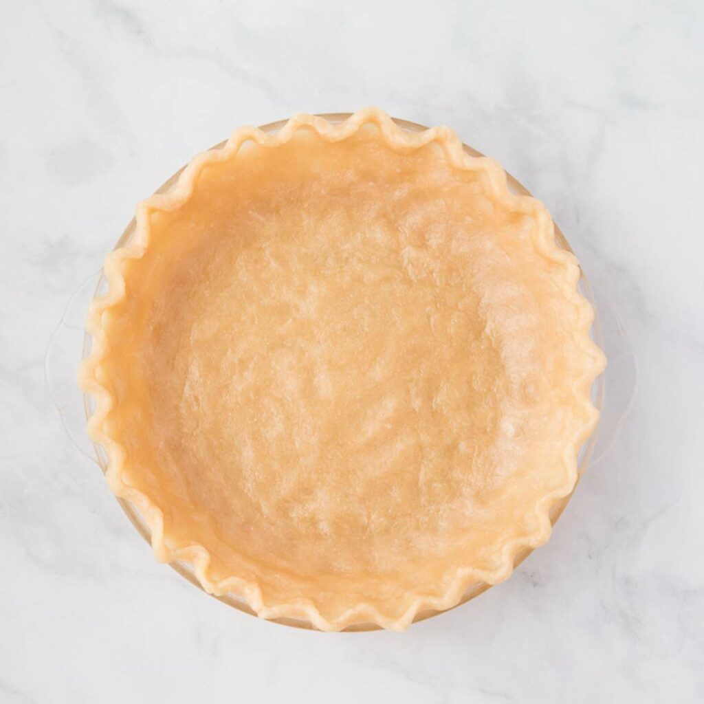 Unbaked pie crust with crimped edges in a glass pie dish on a white marble surface.