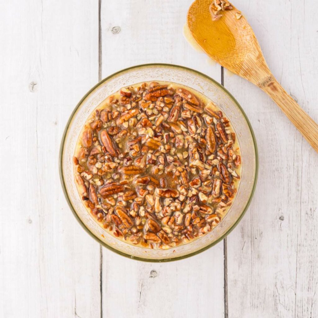 A glass bowl filled with chopped pecans in a brown liquid mixture sits on a white wooden surface next to a wooden spoon.