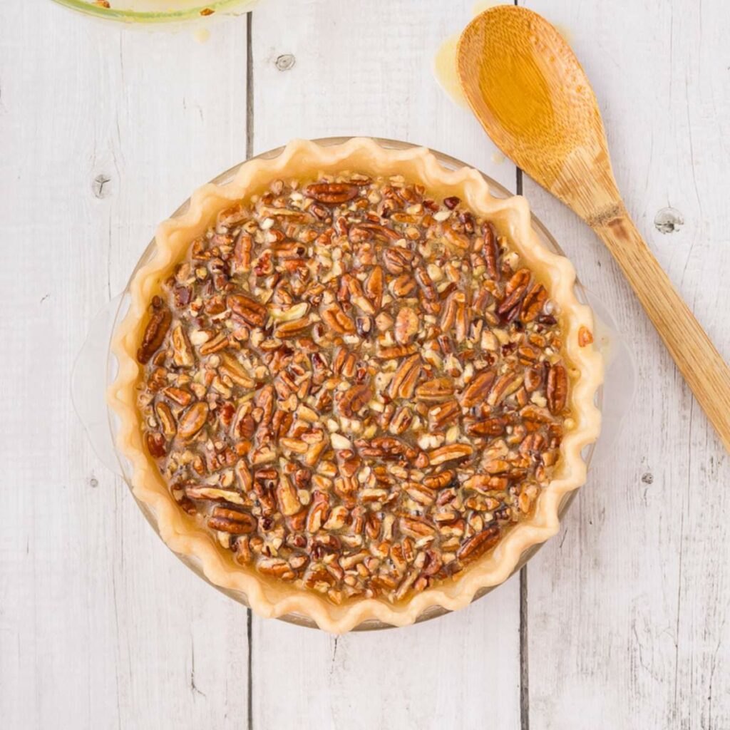 Unbaked pecan pie in a pie dish on a white wooden surface, with a wooden spoon beside it.