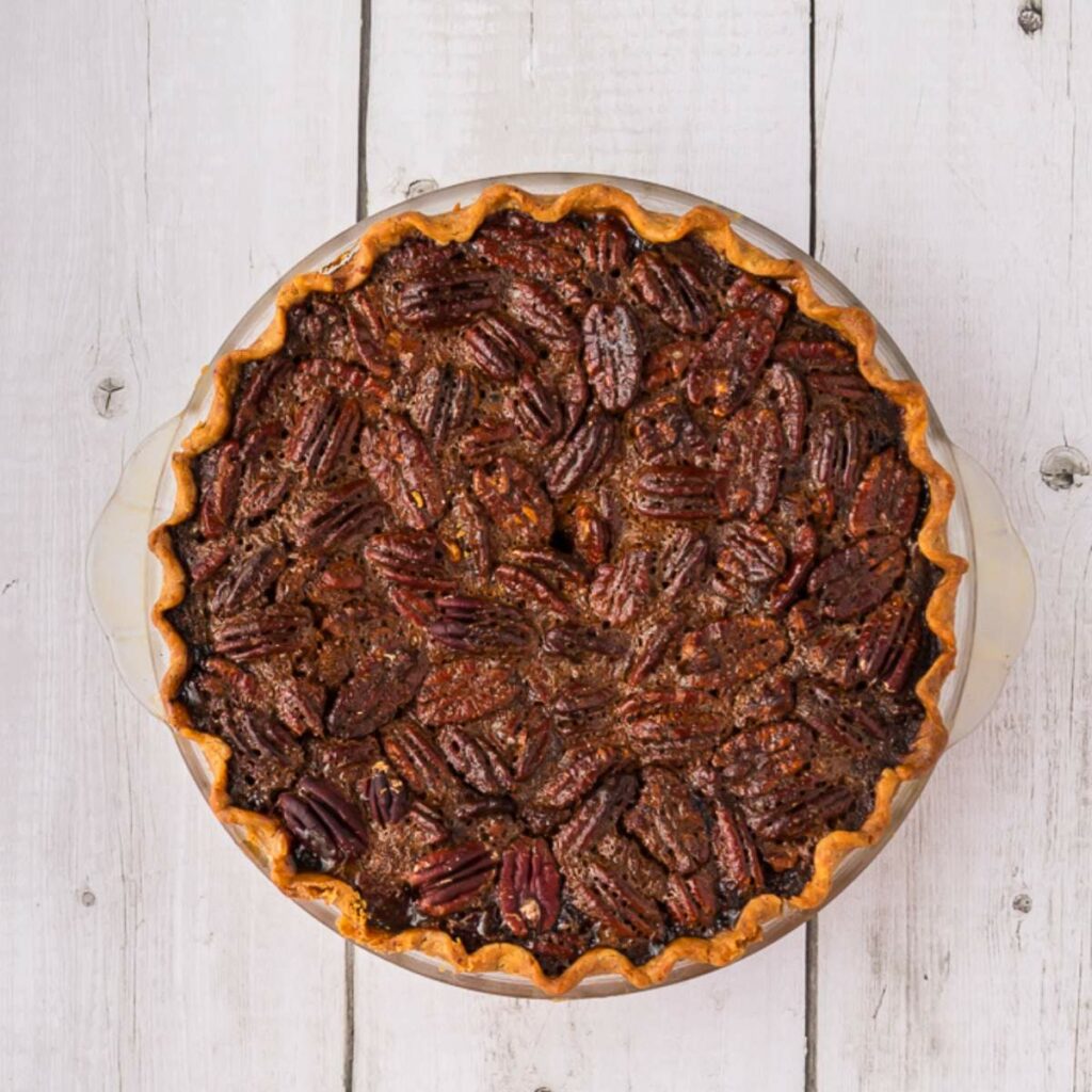 A whole pecan pie with a golden crust, viewed from above, sits on a white wooden surface.