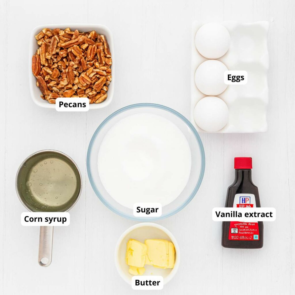 Top-down view of pecans, eggs, sugar, corn syrup, butter, and vanilla extract arranged in separate containers on a white surface.