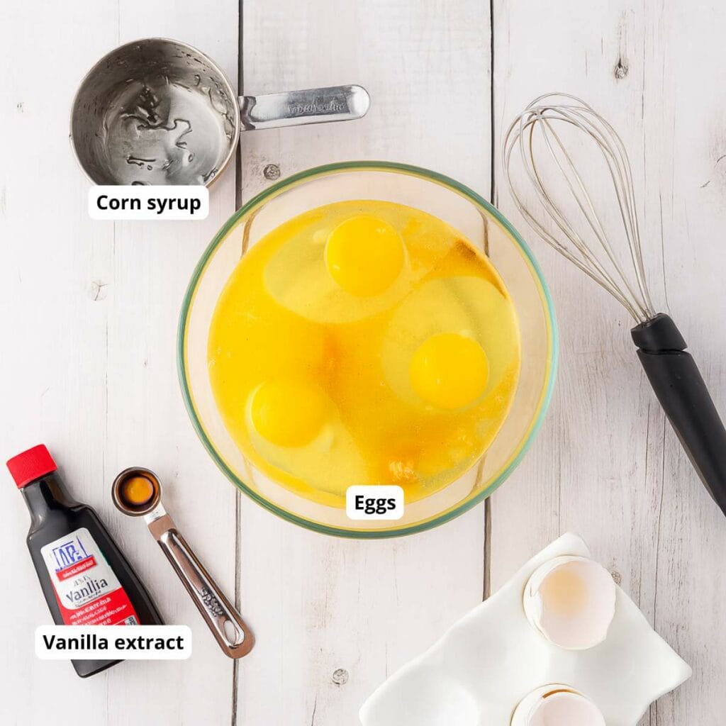 A mixing bowl with eggs, a measuring cup with corn syrup, a bottle of vanilla extract, a whisk, and eggshells on a white wooden surface.
